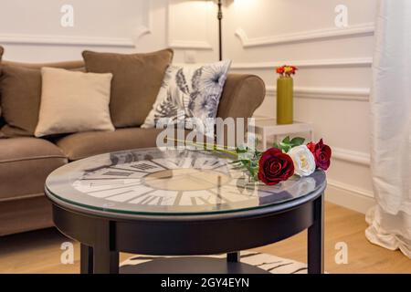 Fresh roses on coffee table with clock inside. Interior of living room with cosy sofa. Stock Photo