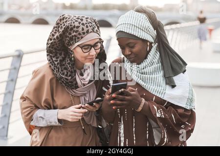 Two friends in muslim clothing discussing photos online on mobile phone during their walk in the city Stock Photo