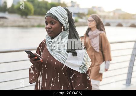 African woman in muslim clothing using her mobile phone while walking along the street Stock Photo