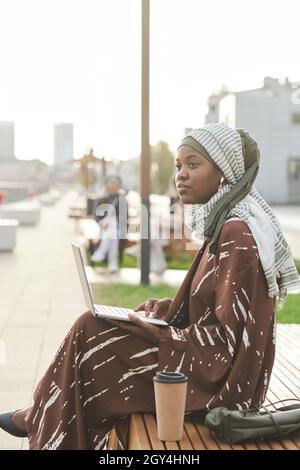 African muslim woman sitting on the bench with laptop and looking awwhile spending time outdoors in the city Stock Photo