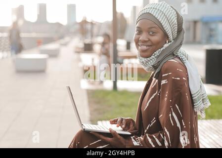 Portrait of African muslim woman smiling at camera while sitting on the bench and using laptop outdoors Stock Photo