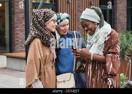 Muslim women talking to their friend while she reading a message on the phone and smiling during their meeting outdoors Stock Photo