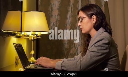 woman in glasses and in a business suit in the evening works at a laptop Stock Photo