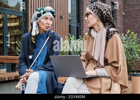 Two business colleagues in muslim clothing using laptop and discussing online work together while sitting on the bench at meeting outdoors Stock Photo