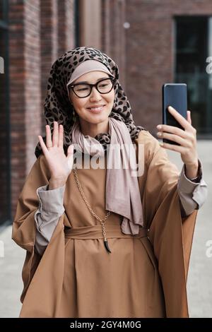 Young blogger in muslim clothing smiling at camera of her phone and talking to her followers while standing outdoors Stock Photo
