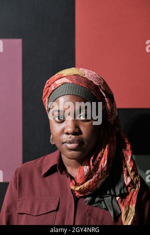 Close-up of African young woman in hijab looking at camera while sitting against the modern wall Stock Photo