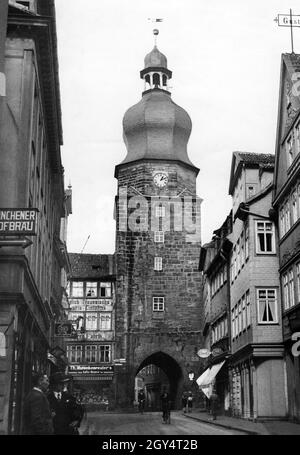 'Two cyclists ride through the Spitaltor into the Spitalgasse in the old town of Coburg. On the sidewalk to the right walks a member of the Sturmabteilung. On the left two men look into the Kleine Johannisgasse, into which the sign above them also points to the ''Münchener Hofbräu''. On the left, across Kleine Mauer past ''Th. Hutschenreuter's Kaffee-Rösterei,'' the path leads to the train station. The undated photo must have been taken before 1937, because in that year additional pedestrian passages were built to the left of the tower. [automated translation]' Stock Photo