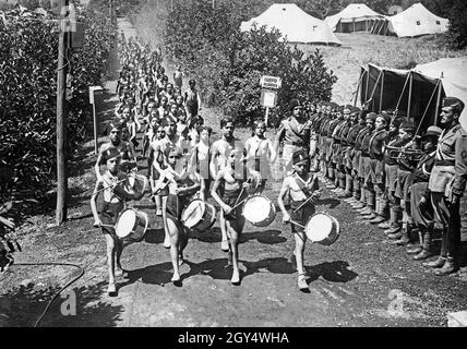 'A group of children and young people, dressed only in shorts, run across a country lane in the summer of 1933, led by four drummers who set the beat and tempo. On the right, another group stands in formation in front of a tent that forms the ''gatehouse'' of the camp (sign: Corpo di Guardia, below which hang the day's orders). The youths are members of the Balilla and are on a holiday camp with sports exercises for ''physical training''. [automated translation]' Stock Photo