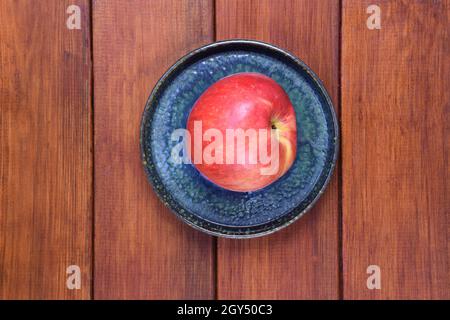 A red apple lies on a ceramic saucer on a wooden brown background. The saucer stands in the center of the frame. Stock Photo
