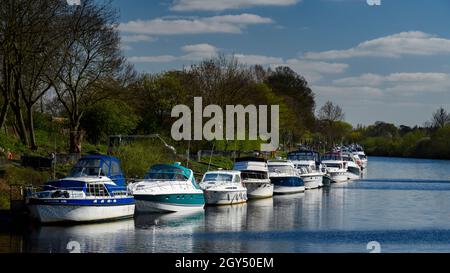 Line of sunlit boats (leisure craft) moored on River Ouse at riverbank moorings on sunny summer day - Fulford, York, North Yorkshire, England, UK. Stock Photo