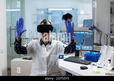 Biologist researcher wearing virtual reality headset examining biochemistry experiment working in microbiology hospital laboratory using biotechnology. Specialist scientist analyzing virus development Stock Photo
