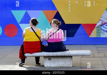 London, England, UK. Two colourful women sitting in front of a colourful wall in Oxford Street on a sunny day in October [no model release] Stock Photo