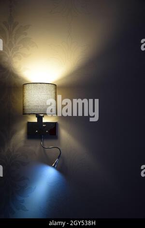 An electric wall lamp hangs on a decorated wall in the twilight. The upper part glows yellow and the lower part glows blue. Stock Photo