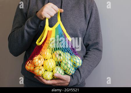 A man holds in his hands a reusable cotton shopping string bag, a multicolored rainbow LGBT with green apples inside. On a gray background. Zero waste Stock Photo