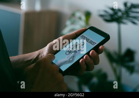 a young caucasian man, at his home, looks for a psychologist online with his smartphone, with a simulated search engine in its screen that reads psych Stock Photo