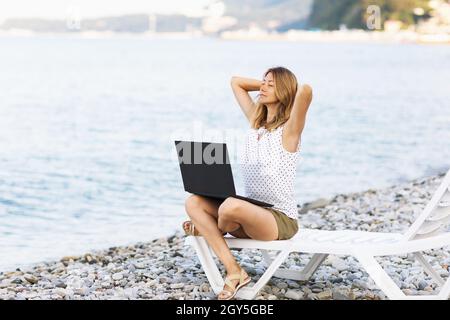 A woman sits on a sun lounger with a laptop on the seashore, combines rest and work Stock Photo