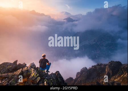 Man sits alone on the peak of rock. Hiker watching to autumn Sun at horizon . Beautiful moment the miracle of nature. Colorful mist in valley. Man hik Stock Photo