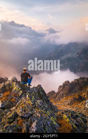 Male hiker takes a break and enjoys mountain views. Female hiker backpacker sitting on the peak edge and enjoying mountains view valley during heavy m Stock Photo