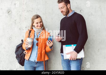 Two students with phone and books standing in front of concrete wall on campus Stock Photo