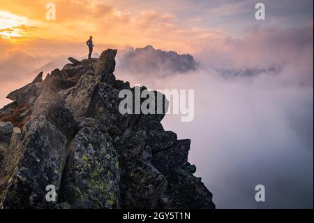 Man stands alone on the peak of rock. Hiker watching to autumn Sun at horizon . Beautiful moment the miracle of nature. Colorful mist in valley. Man h Stock Photo
