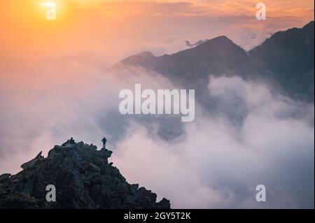 Man stands alone on the peak of rock. Hiker watching to autumn Sun at horizon . Beautiful moment the miracle of nature. Colorful mist in valley. Man h Stock Photo