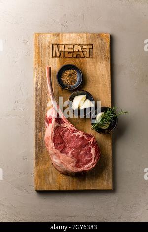 Raw fresh meat Tomahawk Steak and seasonings on wood board on concrete background Stock Photo