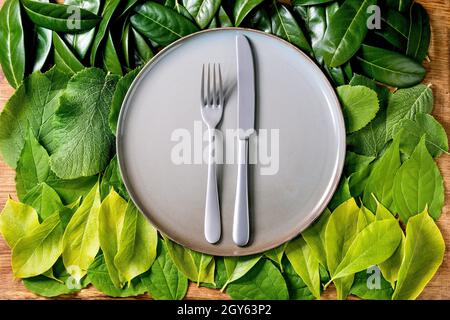 Empty ceramic plate with knife and fork on background made of green leaves, green gradient. Copy space. Empty place for product. Summer menu concept. Stock Photo