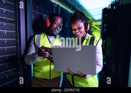 Happy african american computer technicians wearing headphones using laptop working in server room Stock Photo