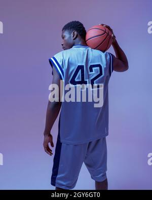 Basketball player poses with ball in studio, back view, neon background. Professional male baller in sportswear playing sport game, tall sportsman Stock Photo