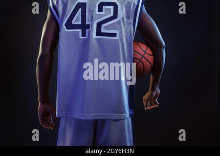 Basketball player poses with ball in studio, back view, black background. Professional male baller in sportswear playing sport game, tall sportsman Stock Photo