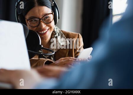 happy asian radio host in headphones using smartphone near blurred colleague typing on laptop Stock Photo