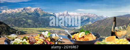 Panorama of Tauer Alps with traditional Austrian specialties such as steaks, fries, potatoes, a bottle of wine against Zell am see town in  Austrian A Stock Photo