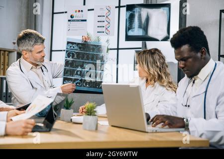 Male and female doctors in white lab coat looking at tomography scan of patient. Group of multiracial colleagues discussing clinical case at office for making diagnosis of severe patient. Stock Photo