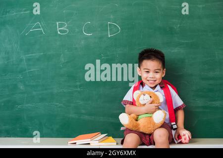 Back to School. Happy Asian funny cute little child boy from kindergarten in student uniform with school bag smiling and hugging teddy bear on green s Stock Photo