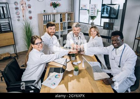 Group of five diverse doctors stacking hands in circle for successful cooperation during meeting. Medical workers with one colleague in wheelchair smiling and looking at camera at office room. Stock Photo