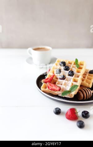 Stack of waffles on a plate on the white table with blueberry, chopped strawberry and mint leaves and white cup and plate of coffee. High quality phot Stock Photo