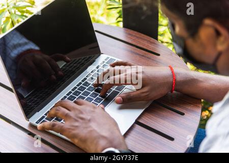 Happy Asian black businessman person or office worker wear face mask protect sitting on desk work from home using laptop computer typing keyboard and Stock Photo