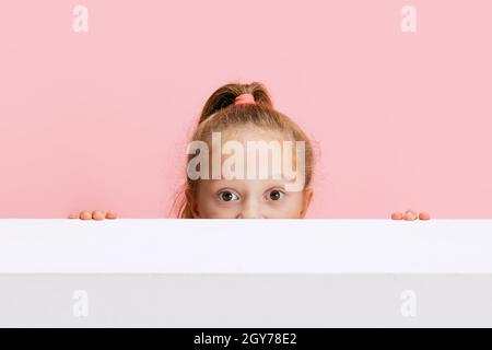 Portrait of funny cute beautiful girl peeping out from behind the wall isolated on pink background. Stock Photo