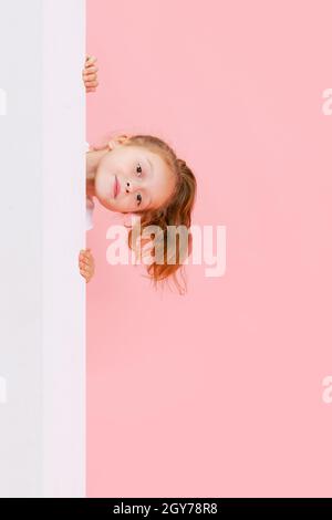 Portrait of funny cute beautiful girl peeping out from behind the wall isolated on pink background. Stock Photo