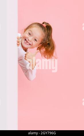 Portrait of funny cute beautiful girl peeping out from behind the wall isolated on pink background. Stock Photo