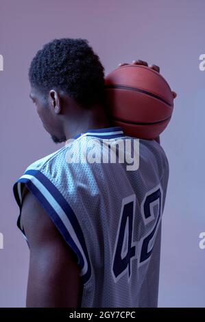 Basketball player poses with ball in studio, back view, neon background. Professional male baller in sportswear playing sport game, tall sportsman Stock Photo