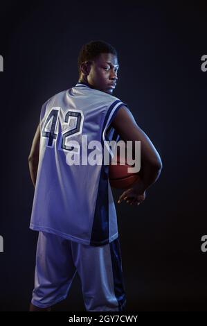 Basketball player poses with ball in studio, back view, black background. Professional male baller in sportswear playing sport game, tall sportsman Stock Photo