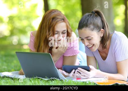 Happy students watching online videos on smart phone laughing and lying on the grass in a park Stock Photo