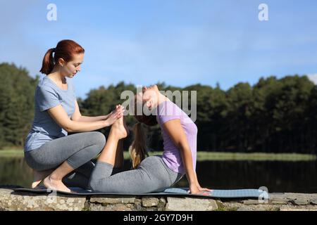 Side view portrait of a woman helping another to do yoga exercise in nature Stock Photo