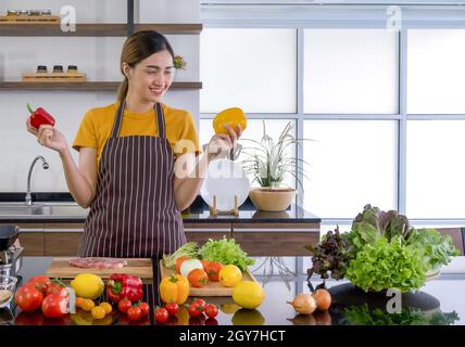 Young housewife stand smiling,  hold red and yellow bell pepper with both hands. Looking at the yellow one on the left. The kitchen counter full of va Stock Photo