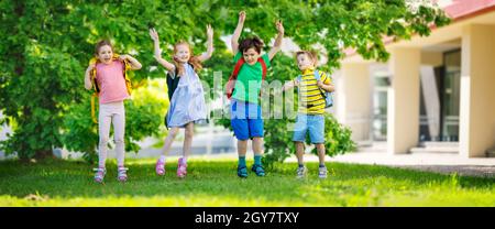 Cute children with rucksacks jumping and playing in the park near the school. Concept of elementary studying and friendship. Stock Photo
