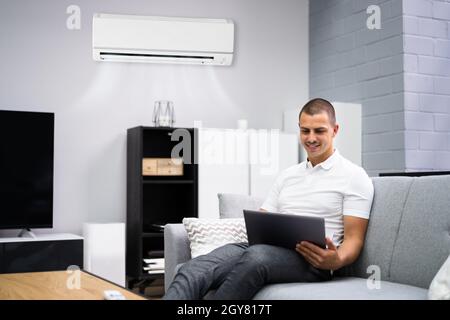 Young Happy Man Sitting On Couch Using Air Conditioner At Home Stock Photo