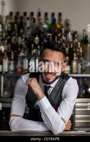 Smiling young bearded male bartender in elegant outfit leaning on bar counter and looking at camera against blurred bottles of alcoholic drinks Stock Photo