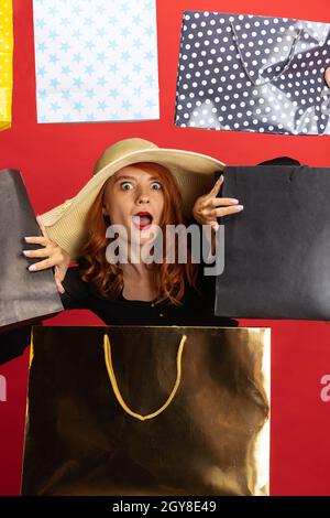 Cropped portrait of beautiful redheaded woman in hat surrounded by many packaging isolated over red background. Holiday shopping Stock Photo