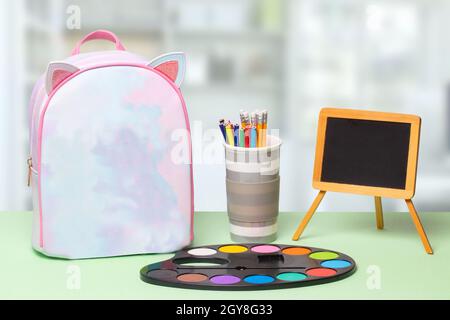 Back to school. Colorful school equipment, a bright school backpack with space for text and a empty blackboard on green table over an abstract bright Stock Photo